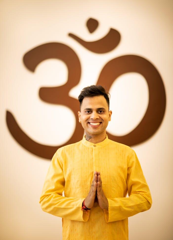 pexels-photo-13849069 A smiling man in traditional yellow attire stands in front of an Om symbol, showcasing spirituality.